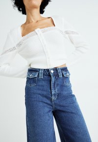 Witte gebreide cropped cardigan met een kantafwerking, voorzien van een knoopsluiting. Gecombineerd met hooggewijde blauwe denim jeans in een relaxed fit.