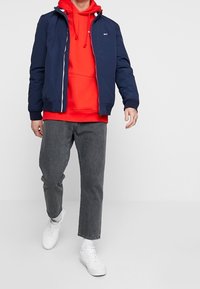 Giacca bomber navy sopra un hoodie rosso, jeans grigi e sneakers bianche. La giacca presenta una cerniera e polsini a coste.