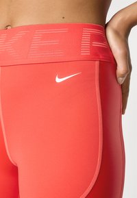 Plan prikaza osobe koja nosi svijetlocrvene Nike Pro tajice s visokim pojasom i bijelim Nike swoosh logotipom na boku.