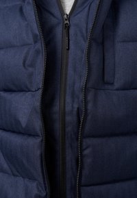Mørkeblå puffvest med herringbone tekstur, der har en central lynlås, sidelomme og en glat sort lynlås.