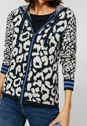 Femme portant un sweat à capuche zippé imprimé léopard noir et blanc avec des poignets rayés bleus et un jean bleu, tenant un objet bleu assorti.
