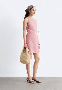Femme debout portant une robe rose sans manches, un foulard à motifs sur la tête, des talons ouverts noirs, tenant un sac tissé beige.