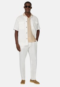 Camicia bianca a maniche corte con due tasche frontali, maglietta beige a scollo a V sotto, pantaloni bianchi e scarpe slip-on marrone chiaro.