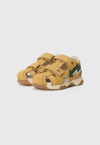 Sandales en cuir jaune avec des côtés ouverts, deux bandes velcro, coutures blanches et un accent vert sur le côté. Semelle en caoutchouc pour une meilleure adhérence.