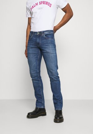 Hombre vestido con jeans ajustados azules, botas negras y una camiseta blanca con el texto "PALM SPRINGS", de pie contra un fondo liso.