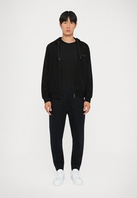 Svart hettejakke med teksturert design, kombinert med en svart t-skjorte og marineblå joggebukser. Hvite sneakers fullfører antrekket.