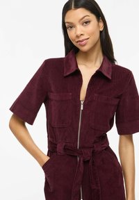 Tuta in velluto a coste bordeaux con maniche corte, cerniera frontale, due tasche sul petto e dettaglio in vita con cintura annodata. La texture appare morbida e a coste.
