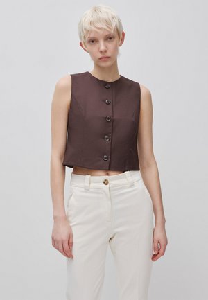 Junge Person mit kurzen blonden Haaren, trägt ein ärmelloses dunkelbraunes, geknöpftes Oberteil und hoch taillierte weiße Hosen vor schlichtem Hintergrund.