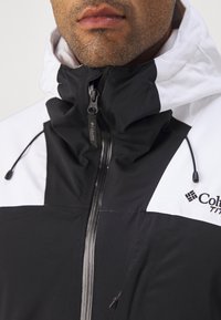Juoda ir balta vandeniui nepralaidi striukė su aukštu apykakle, priekiniais užtrauktukais ir reguliuojama kapuce. Yra Columbia logotipas ir tekstūruotas audinys.