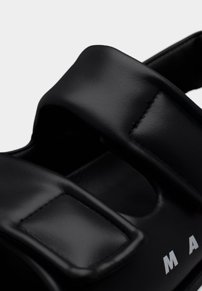 Μαύρα σανδάλια με δύο ρυθμιζόμενους ιμάντες από λείο υλικό, με ραμμένη διακόσμηση και έντονη λευκή επιγραφή στο πλάι.