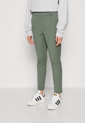 Grüne maßgeschneiderte Hose mit schmaler Passform und flachem Frontdesign. Kombiniert mit weißen Sneakers mit schwarzen Streifen und einem grauen Sweatshirt.