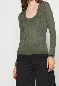 Haut à manches longues côtelé vert olive avec un col rond et une texture scintillante. Coupe slim, avec un petit détail de marque sur l'ourlet.