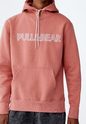 Personne portant un sweat à capuche rose poudré avec le texte blanc "PULL&BEAR", une poche avant et des cordons, associé à un pantalon gris foncé.