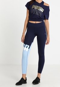 Námorně modré zkrácené tričko s zlatým logem "PUMA", spárované s námořnickými a světle modrými legínami s bočním logem. Černé sportovní boty.