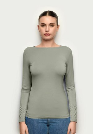 Kobieta o brązowych włosach związanych do tyłu, ubrana w długi rękaw szarego topu i niebieskie dżinsy, stoi na tle jednolitego białego tła.