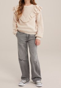 Krémový pletený svetr s volánkovým detailním výstřihem, v kombinaci se zašedlými džínami s místy s opotřebením a bílými botami s leopardím vzorem.