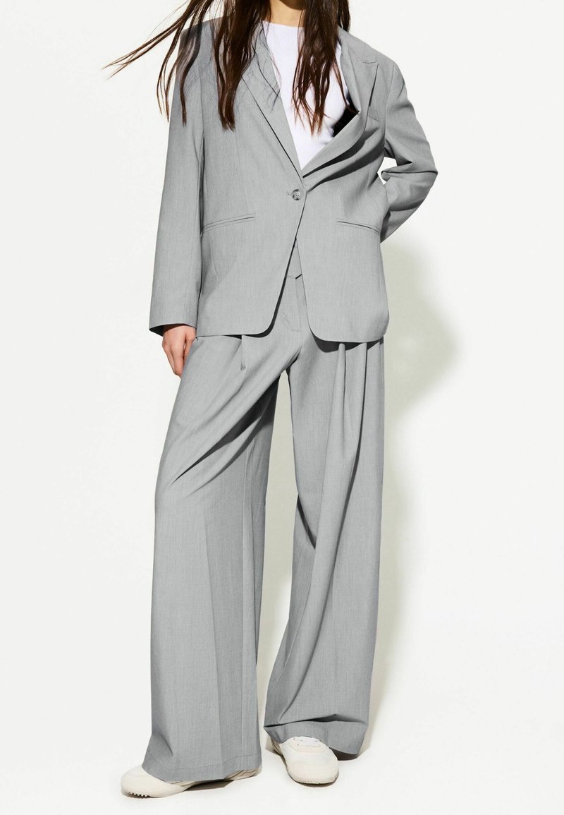 Femme portant un costume gris clair ample avec un pantalon large, une chemise blanche et des baskets blanches, debout devant un fond uni.