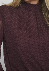 Blusa borgogna con collo alto arricciato, caratterizzata da un motivo a zigzag testurizzato e maniche lunghe trasparenti.