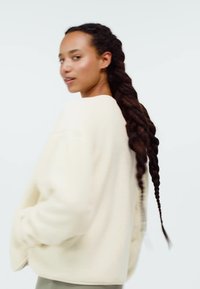 Ung kvinna med långt flätat hår som bär en lös off-white tröja, vänd lätt åt sidan mot en enfärgad bakgrund.