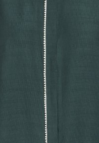 Tela verde oscuro con un acabado texturizado, que presenta una costura con cremallera plateada adornada con pequeñas cuentas blancas a lo largo del borde.