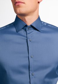 Lichtblauwe, lange mouwen shirt met een puntkraag, een gladde textuur en een knoopsluiting. Beschikt over parelmoeren knopen als accenten.