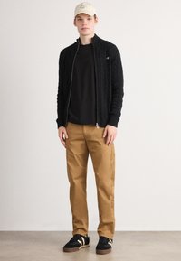 Ung mand med en beige kasket, sort lynlås kabelstrikket sweater, sort t-shirt, lysebrune bukser og sorte sneakers med beige striber står på en grå bund.