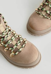 Stivali da escursionismo in suede beige con lacci a motivo verde e marrone e dettagli in oro. Suola in gomma spessa con superficie testurizzata. Design della punta angolare.