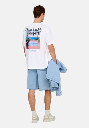 Junger Mann trägt ein weißes T-Shirt mit Tennisplatzmuster und Text, Jeansshorts, weiße Turnschuhe, hält eine leichte Jeansjacke in der Hand.