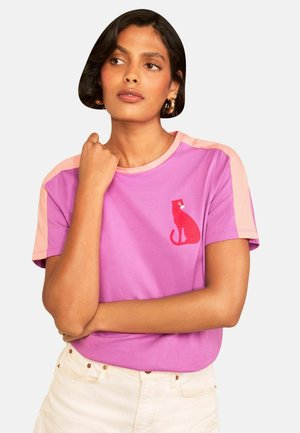 Frau mit kurzen dunklen Haaren trägt ein pink-lila Hemd mit einer roten Katzengrafik auf der Brust und hellen Hosen.