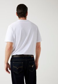 Hvid kortærmet t-shirt set fra ryggen, parret med mørkeblå denim jeans med gul syning og et logo på baglommen.