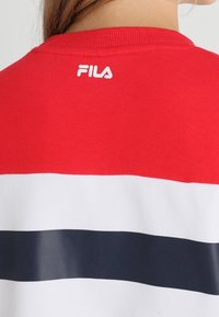Sudadera roja con rayas blancas y marinas. Cuenta con un cuello acanalado y el logo de FILA en blanco en la parte posterior, cerca del cuello.