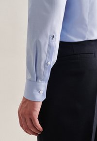 Camicia azzurro chiaro con polsini bottonati, caratterizzata da una forma esagonale e dettagli ricamati di fiori. Tessuto liscio, abbinata a pantaloni scuri.
