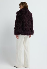 Person iført en mørk lilla tekstureret pelsjakke, hvide vide bukser og briller, der står foran en ensfarvet lys baggrund.