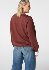 Bruine trui met lange, opgeblazen mouwen en geribbelde zoom, gecombineerd met een high-waisted blauwe spijkerbroek. Witte kanten accenten aan de mouwboorden.