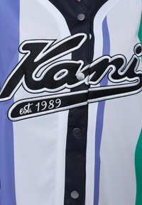 Beisbola stila krekls baltā, tumši zila un lavandas krāsā. Iekļauj izšūtus uzrakstus, spiedpoges un teksturētu audumu. Horizontālas krāsu joslas.