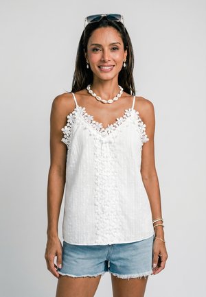 Femme souriante portant un haut blanc à fines bretelles orné de dentelle, un short en jean, un collier de coquillages, des boucles d’oreilles, des bracelets et des lunettes de soleil sur la tête.