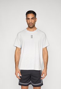 Αθλητική κοντομάνικη μπλούζα ανοιχτού γκρι με διακριτικό μοτίβο διαβάθμισης, λαιμόκοψη τύπου crew και κοντά μανίκια. Συνδυάζεται με μαύρο σορτς από δίχτυ με λευκές λεπτομέρειες.