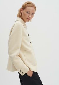 Giacca in misto lana color avorio con colletto, tasca frontale e bottoni neri sulle maniche. Tessuto liscio, forma leggermente oversize, design minimalista.