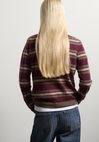 Maglione a strisce bordeaux con linee beige e marroni, orlo a coste, abbinato a jeans in denim blu. Lunghi capelli biondi lisci scendono sulla schiena.