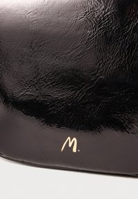 Borsa in pelle nera con una texture lucida, caratterizzata da un logo "M." in rilievo dorato. Superficie liscia con sottili riflessi di luce.