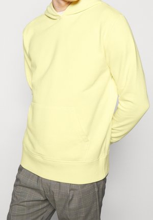 Hombre con sudadera amarilla pálida con capucha y bolsillo frontal, pantalones de cuadros grises, manos detrás de la espalda, mirando ligeramente hacia la izquierda.