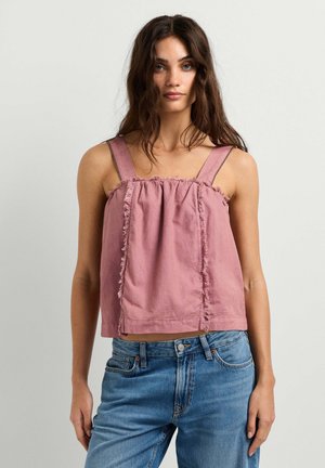 Vrouw met lang donker haar die een mouwloze roze top met ruches en een blauwe spijkerbroek draagt, staand tegen een eenvoudige lichtgrijze achtergrond.