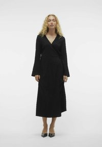 Zwarte wikkeljurk met lange bell mouwen, voorzien van een gestructureerd stippenpatroon. Vloerlange jurk met een V-hals en strikdetail in de taille.