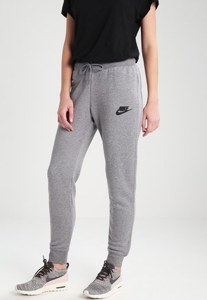 Osoba nosząca szare spodnie dresowe Nike ze ściągaczem w talii i czarnym logo, w parze z czarnym topem i szarymi butami sportowymi.