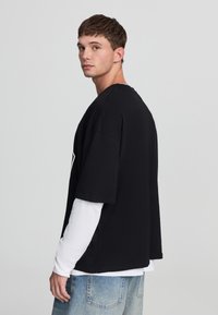 T-shirt nera oversize con maniche corte, indossata sopra una maglietta bianca a maniche lunghe. Abbinata a jeans in denim blu, con una vestibilità comoda.