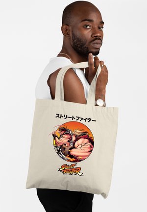 Mężczyzna niesie beżową torbę na ramię z postaciami ze Street Fighter i japońskim tekstem na białym tle, ubrany w białą bluzkę bez rękawów.