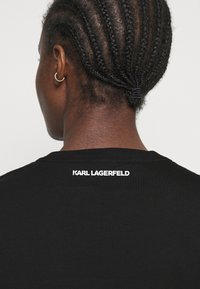 Černé bavlněné tričko s kulatým výstřihem a krátkými rukávy. Na zádech poblíž výstřihu je bílé textové logo "KARL LAGERFELD".