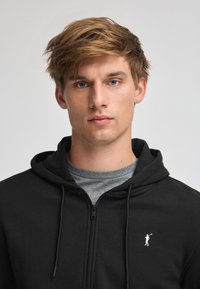 Sudadera con cremallera negra, con cordones, de textura suave, que presenta un pequeño logo blanco de una figura con un palo de golf en el pecho izquierdo.