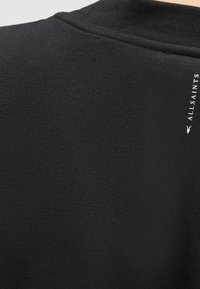 Tissu noir en gros plan montrant un petit logo de marque blanc vertical "AllSaints" près de la couture.