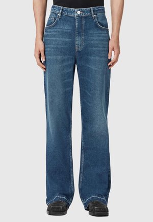 Niebieskie spodnie dżinsowe o prostych nogawkach z przetarciami, noszone z czarnymi butami i czarną górą, pokazane od talii do stóp na jasnym tle.